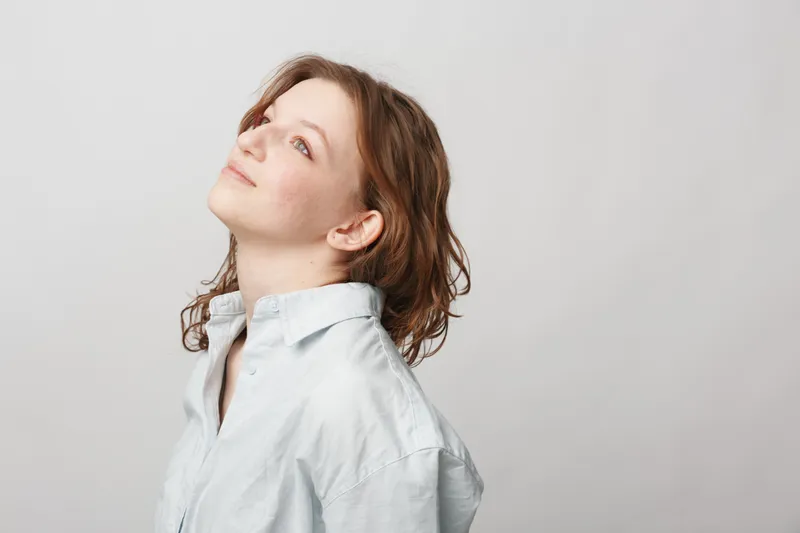 Young woman with short brown hair wearing a white shirt looks upward with a calm expression on a plain light background. Concept of hope, contemplation, and natural beauty.