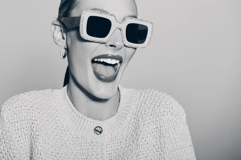 Happy white woman with pale skin wearing large white sunglasses laughs joyfully in studio with light background.