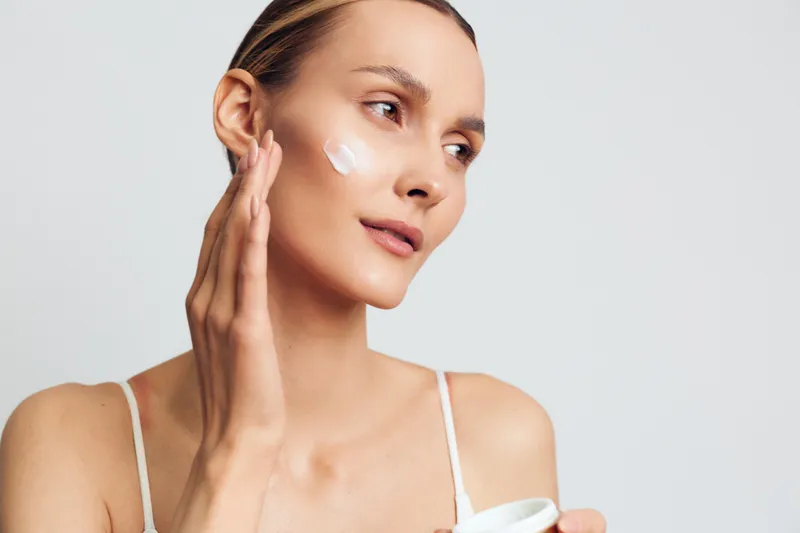 Young woman with clear skin applying white face cream on her cheek. Close-up portrait with soft natural light on neutral background. Concept of skincare, beauty, and daily routine.