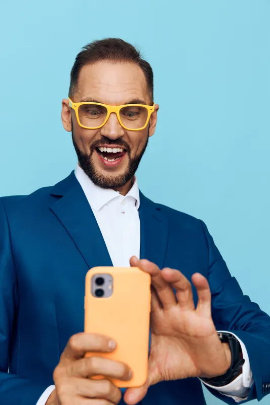 A confident business professional stands against a bright blue background, radiating enthusiasm and focus while capturing a candid moment on a smartphone.
