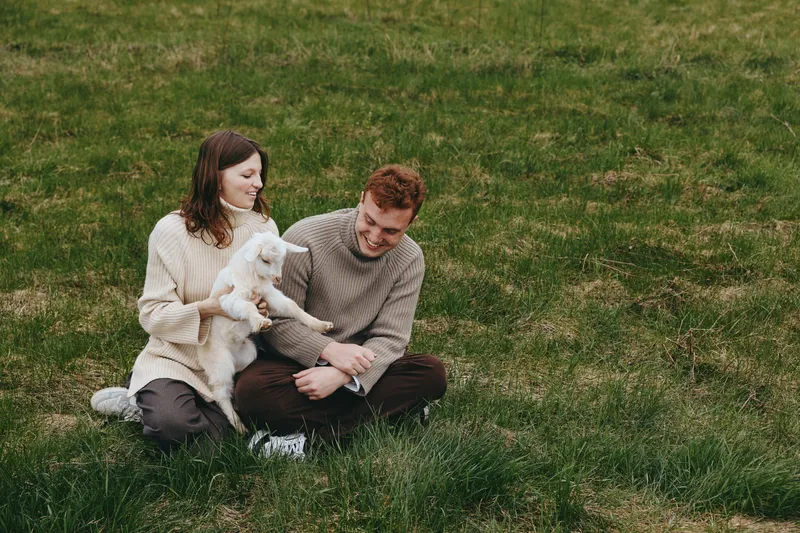 Cheerful couple outdoors, sitting on grass, gentle goat kid nearby, warm sunlight, relaxed moment, candid interaction, soft breeze, natural colors, intimate connection