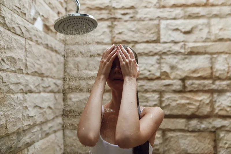 woman washing face under indoor shower, natural stone walls, water droplets, morning hygiene routine, wellness and relaxation atmosphere