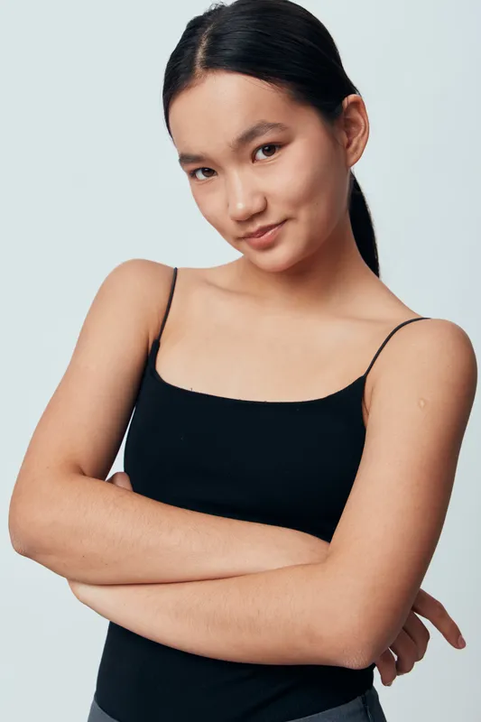 Young woman with long dark hair in black tank top posing with crossed arms and slight smile against plain light background. Natural makeup and confident expression in studio portrait.