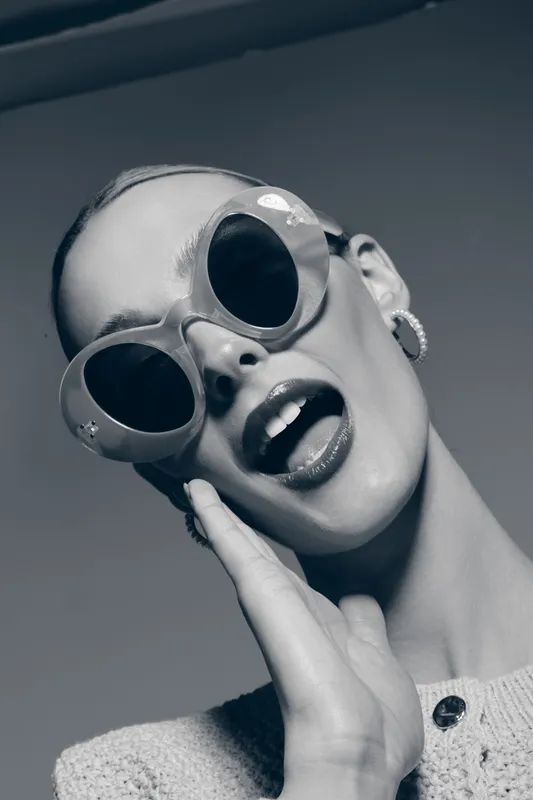 Stylish Caucasian woman with pale skin wearing round sunglasses and earrings poses with open mouth and hand on cheek.