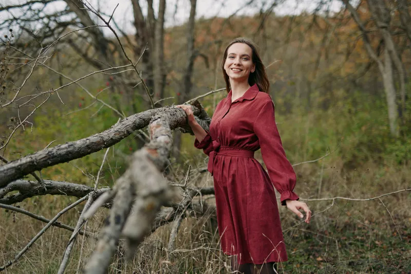 An authentic, candid portrait of a woman in a red dress leaning beside a fallen tree in a calm countryside setting, conveying natural light and credible mood.