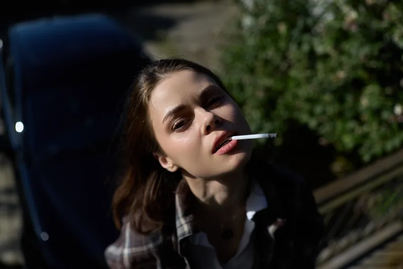 Young woman smoking outdoors leans against a car in a sunlit moment, wearing a plaid shirt with a relaxed mood and moody urban backdrop