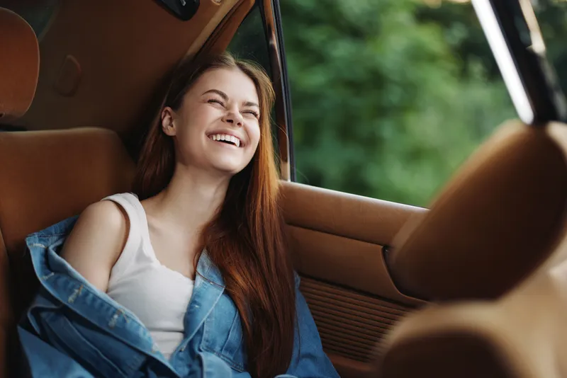 Cheerful young woman with long hair sitting in a car, smiling joyfully, wearing a denim jacket, surrounded by lush greenery, capturing a sense of adventure and freedom.