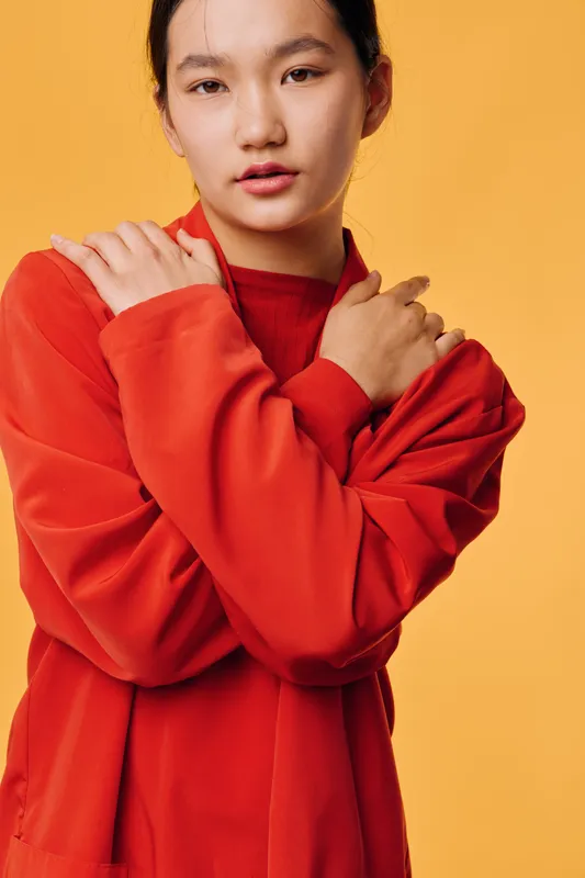 Young woman with crossed arms wearing red sweater posing against yellow background, looking at camera with calm expression and minimal makeup in studio portrait photo.