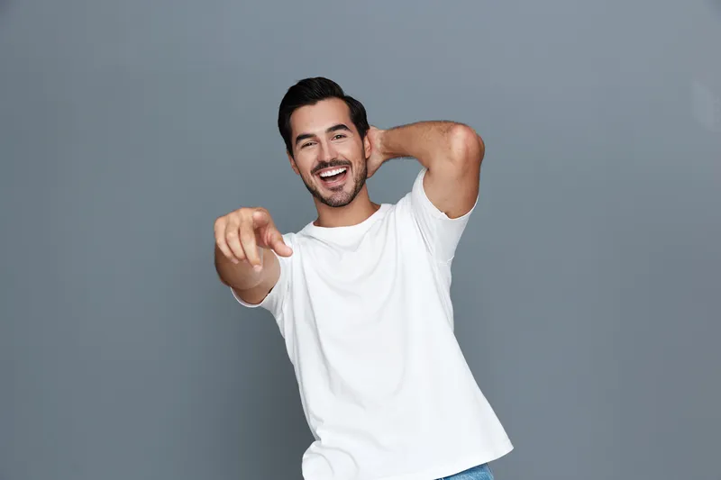 Joyful smiling young man wearing a white t-shirt, leaning against a gray wall while playfully pointing at the camera, showcasing a cheerful attitude and positive energy.
