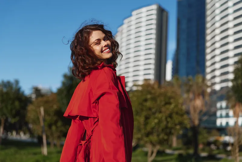 A smiling woman in a bright red coat stands in a city park, sunlight on her face, with tall buildings in the background, conveying confident urban style and optimism.