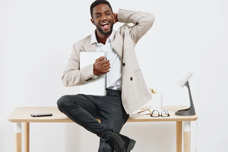 Smiling man sitting on desk holding tablet device in modern office space with minimalist white background. Casual professional pose, happy expression, relaxed atmosphere, contemporary workplace, techn
