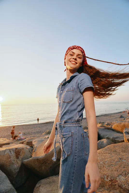 Smiling woman in denim outfit and red headscarf enjoying sunset on rocky beach with flowing hair and warm golden light