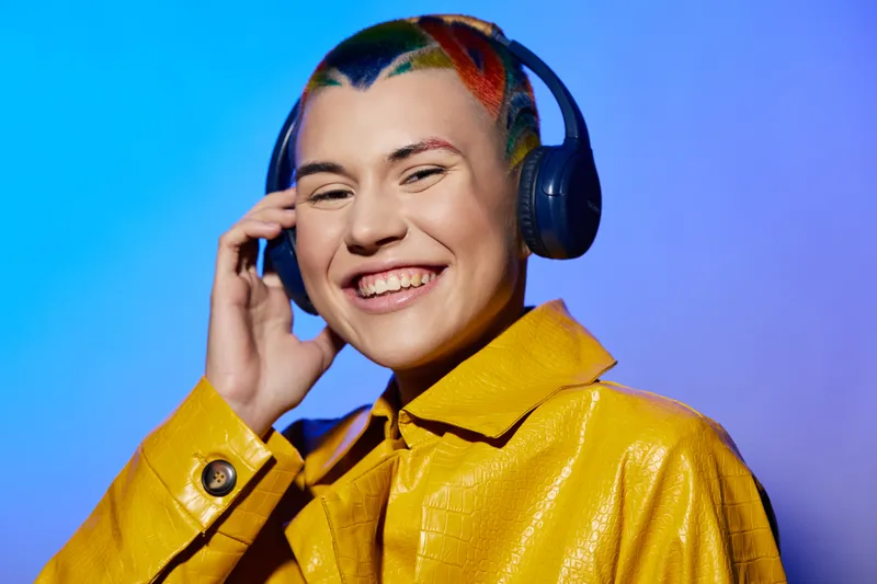 smiling woman with colorful hair wearing headphones and yellow jacket, enjoying music with bright blue background, joyful and vibrant portrait