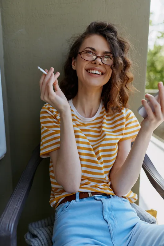 Relaxed woman on a balcony enjoys a casual moment, wearing striped shirt and jeans, smiling with glasses, holding a cigarette, friendly vibe, home lifestyle scene in soft natural light.
