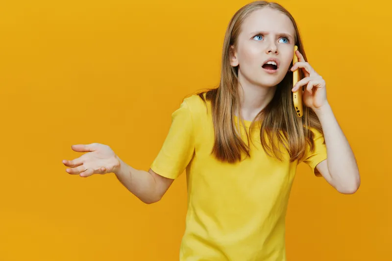 Frustrated girl speaking on the phone against a vibrant yellow background in a casual yellow t shirt, expressing emotions of confusion and annoyance