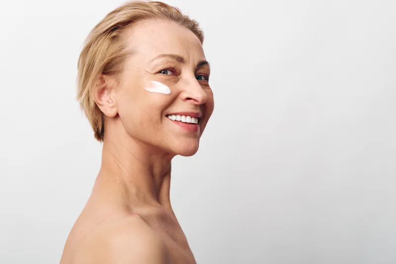 Smiling middle aged woman with short blonde hair applying white face cream on cheek. Close up portrait on light background. Concept of skincare, beauty, wellness, and self care.