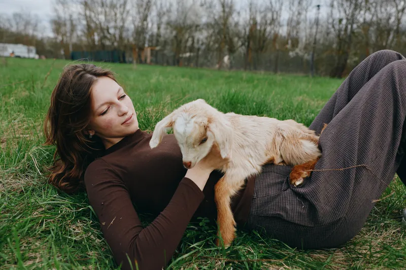 Young woman in a brown turtleneck lies on the grass holding a white baby goat in a peaceful outdoor setting during early spring.