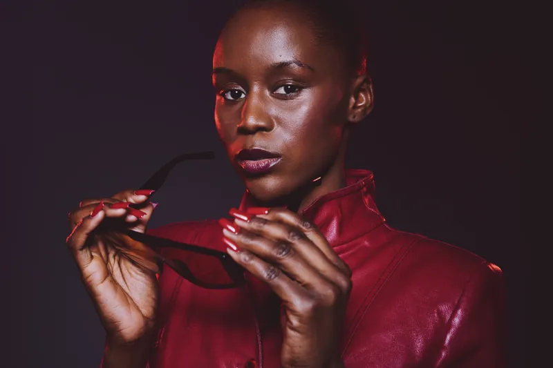 Confident African woman with dark skin wearing a red leather jacket holds sunglasses, posing against a dark background. Concept of fashion, style, beauty, and empowerment.