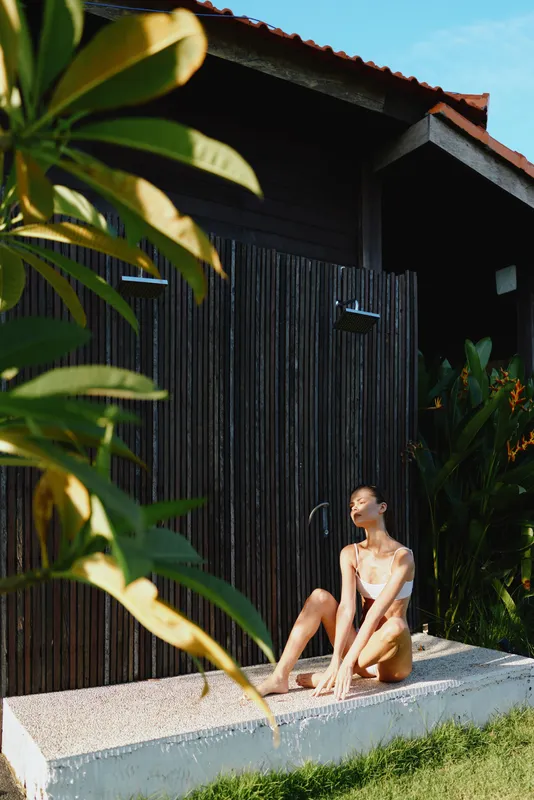 A woman in swimwear relaxes outdoors on a sunny day near a wooden structure surrounded by tropical plants, enjoying a peaceful moment in a natural lifestyle setting.