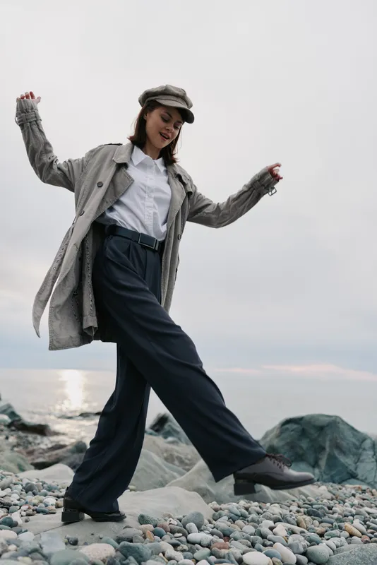 Young woman in casual autumn outfit walks on rocky beach under cloudy sky. She wears coat, trousers, and hat, enjoying nature and fresh air by the sea. Concept for fashion and lifestyle.