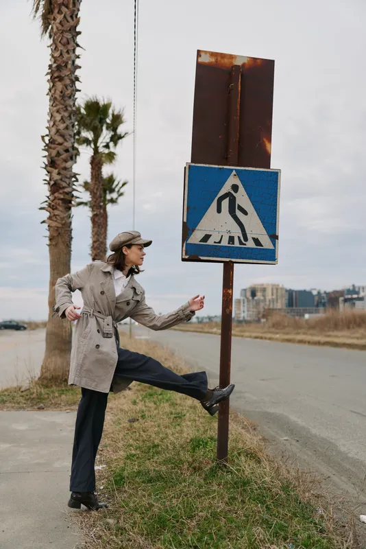A woman in a trench coat and cap playfully poses near a pedestrian crossing sign on a roadside with palm trees and buildings in the background. Urban outdoor concept.