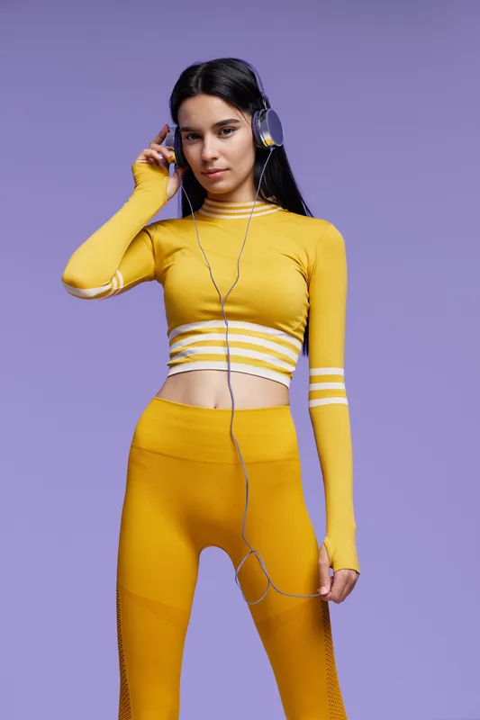 Woman sport fitness wearing yellow athletic outfit and headphones, standing against purple background with focused and relaxed expression, showcasing modern active lifestyle.