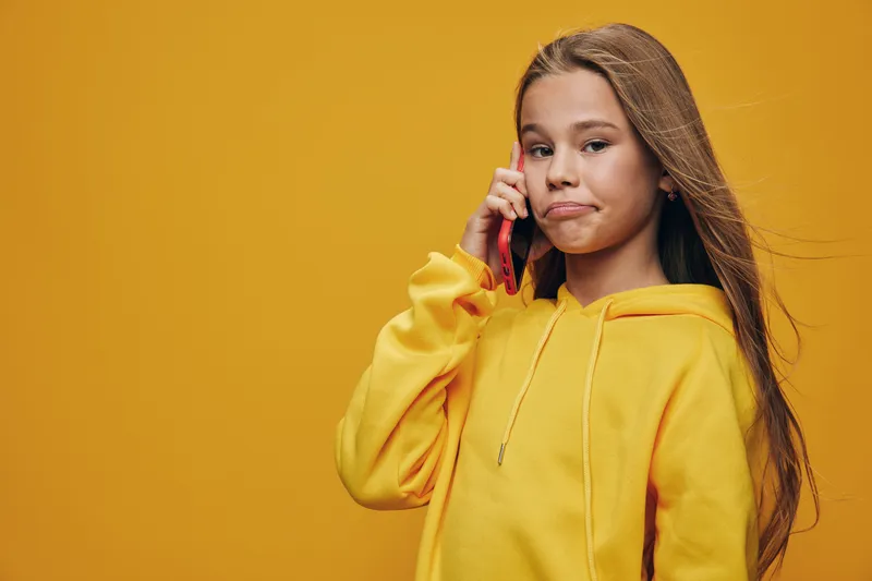 Girl with long hair wearing yellow hoodie talking on smartphone with unsure expression isolated on yellow background. Teenager using mobile phone for communication.