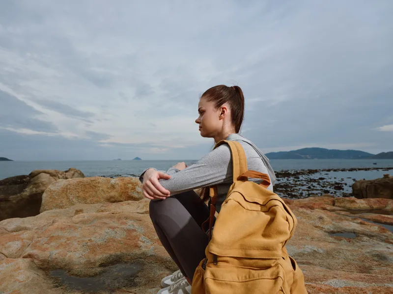 Young woman with backpack sitting on rocky shore, feeling contemplative by the sea, showcasing adventure and exploration in nature s beautiful environment