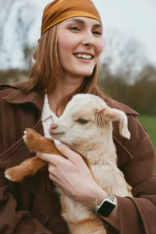 Cheerful woman outdoors cuddling a goat kid, warm sunlight, relaxed pose, rustic field backdrop, casual jacket, soft breeze, wholesome lifestyle moment in the countryside, close interaction, gentle