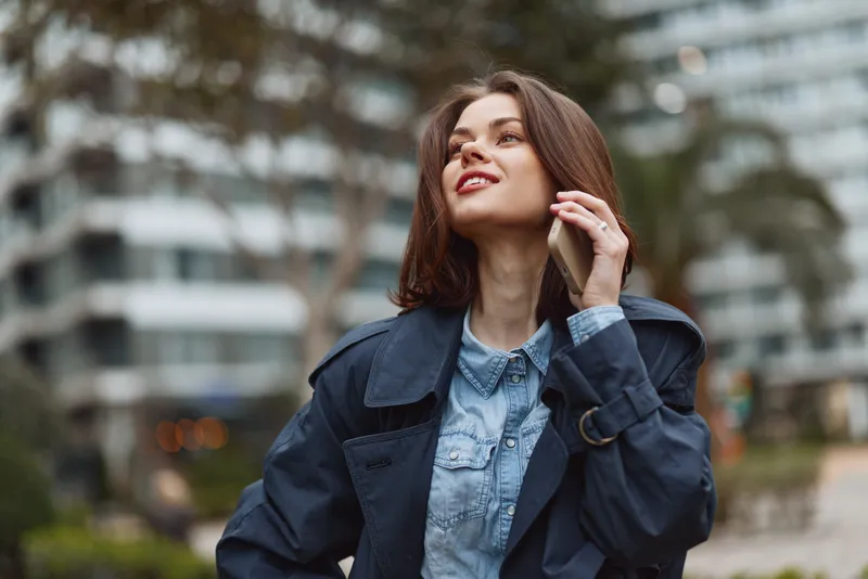 Confident young woman in a denim jacket chats on her phone in a city park, enjoying a casual outdoor moment with a bright smile, background suggesting urban life.
