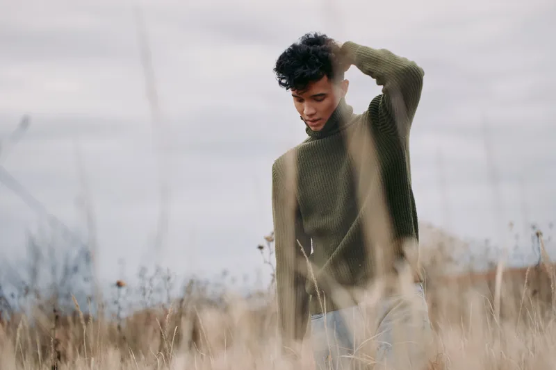 A young man in a knit sweater stands amid tall grasses, looking down with a pensive expression and soft natural light, a candid outdoor portrait conveying authenticity and credibility.