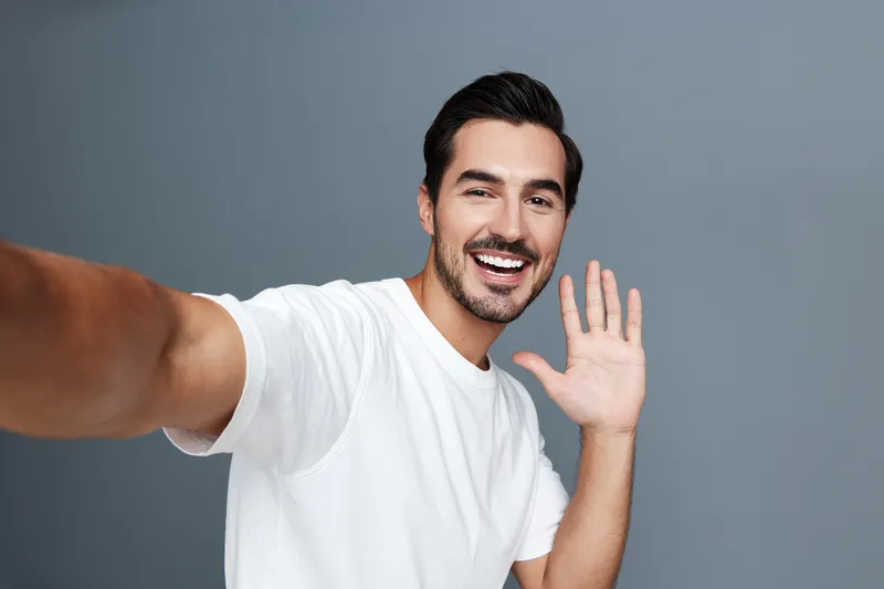 Happy smiling young man waving at the camera while taking a selfie, dressed in a casual white t-shirt, against a solid grey background. People lifestyle concept.