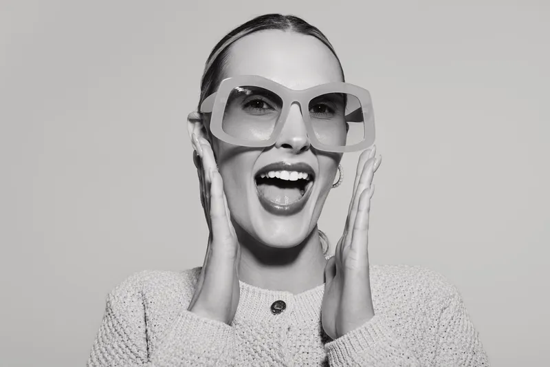 Happy white woman with light skin and pulled back hair wears oversized glasses and knitted sweater, smiling with open mouth on grey background. Concept of joy and fashion.