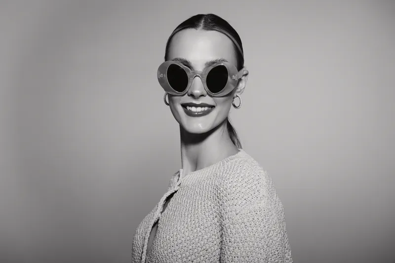 A smiling Caucasian light skin woman with slicked back hair wears round sunglasses and hoop earrings. She poses confidently in studio with soft lighting and grey background.
