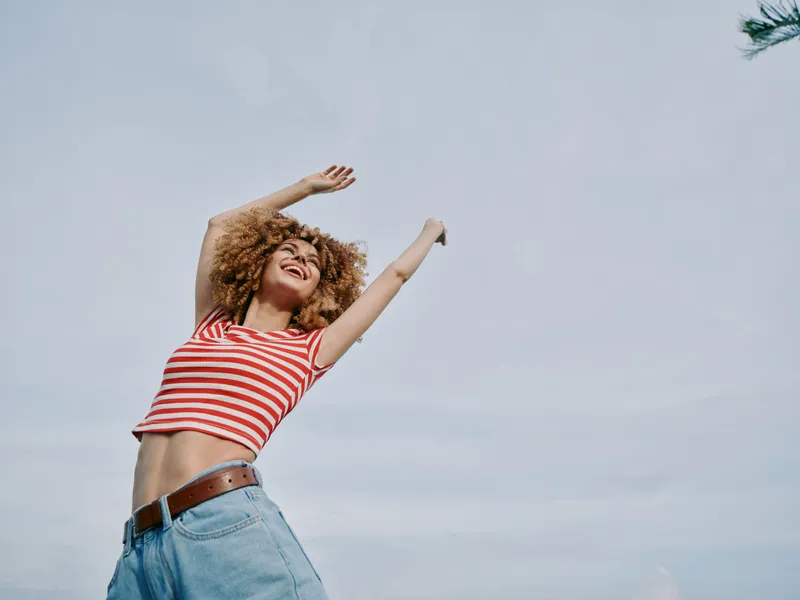 Joyful young woman leaps with arms raised in a sunlit outdoor setting, wearing a red and white striped top and denim jeans, capturing lively motion and carefree energy.