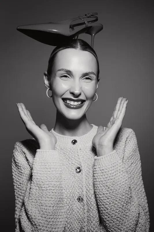 Happy young caucasian woman with light skin smiling broadly wears high heel shoe on head and knitted cardigan. Studio portrait with playful mood and creative fashion concept.