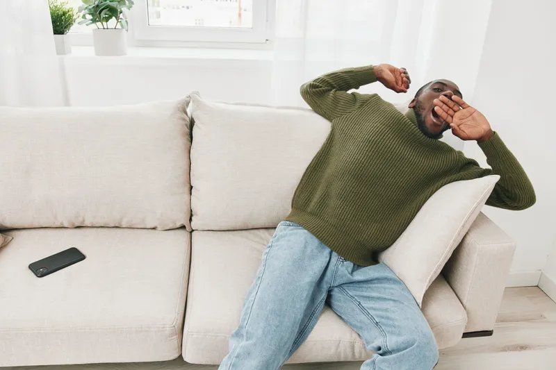 A young Black man in a green sweater and blue jeans yawning on a cozy white sofa, showcasing relaxation and comfort at home during a sunny season