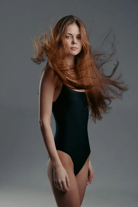 Young woman with flowing hair in black swimsuit posing against gray background, elegant and confident expression