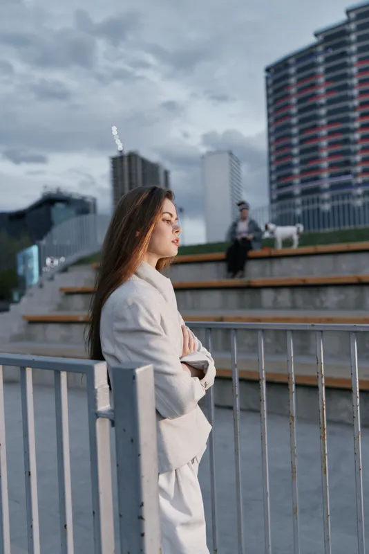 Woman in neutral coat standing thoughtfully near metal fence in urban setting with modern buildings and cloudy sky, conveying introspective mood and fashion style