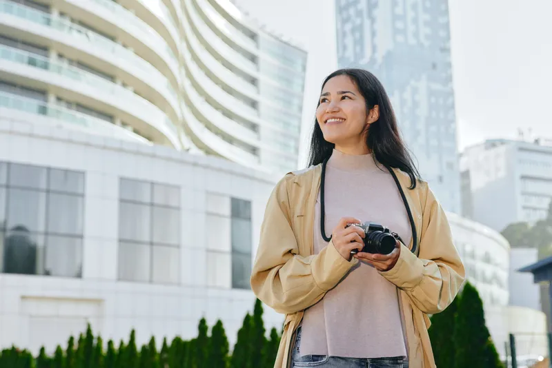 Woman with camera stands outdoors in an urban cityscape, smiling while ready to shoot, wearing a beige jacket and soft sweater, modern buildings visible in the background, conveying travel,