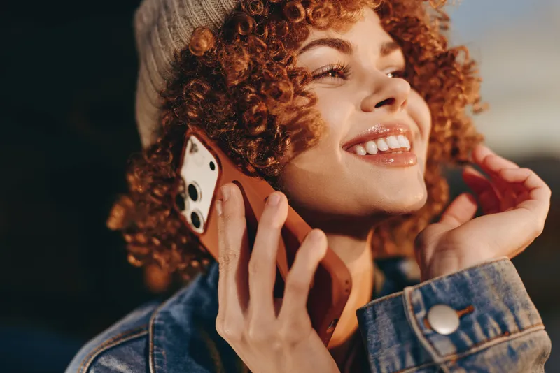 Woman lifestyle curls smartphone smile call denim outdoor, cheerful young woman with curly hair holds smartphone to ear, denim jacket, warm sunlight, soft bokeh background, candid portrait