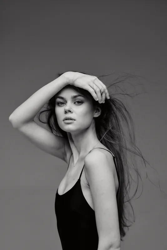 Black and white portrait of a young woman with flowing hair wearing a black top, posing with her hand on her forehead.