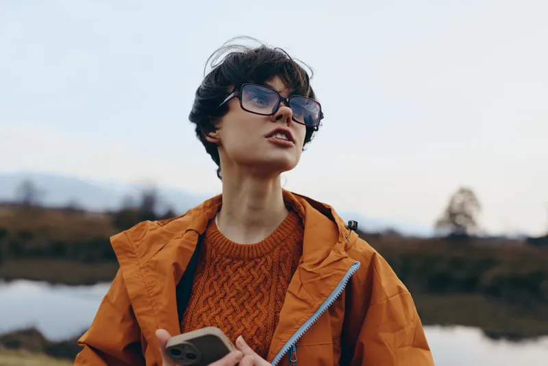 Woman lifestyle outdoors lake smartphone glasses orange jacket, standing near water at sunset, calm expression, casual travel portrait, holding phone with both hands