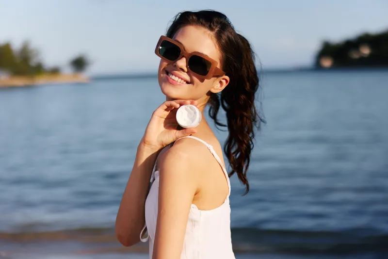 Smiling woman in sunglasses holding sunscreen by the water enjoying summer outdoors, sun protection, vacation, relaxed mood, bright sunny day.