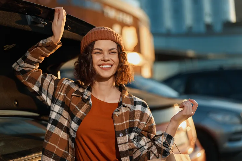 Woman smiling by car trunk with shopping bags in golden hour light, candid lifestyle portrait of urban leisure emphasizing authenticity, golden hour glow, mindful living and emotional storytelling.