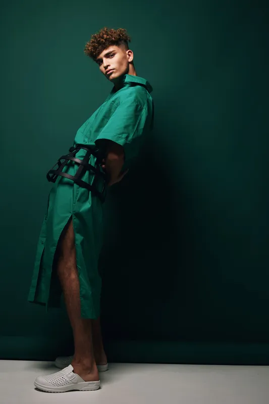 Young man posing in a green outfit with black harness detail and white slipon shoes studio portrait with moody lighting and dark green background modern fashion concept.