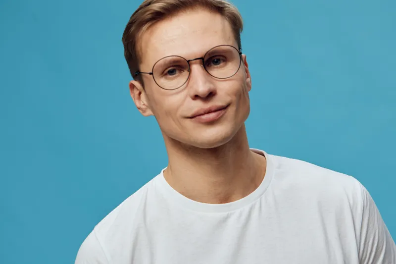 Close-up portrait of a young man with short brown hair, wearing glasses and a white t-shirt, looking confidently at the camera against a bright blue background. Studio shot, casual style, human face, 