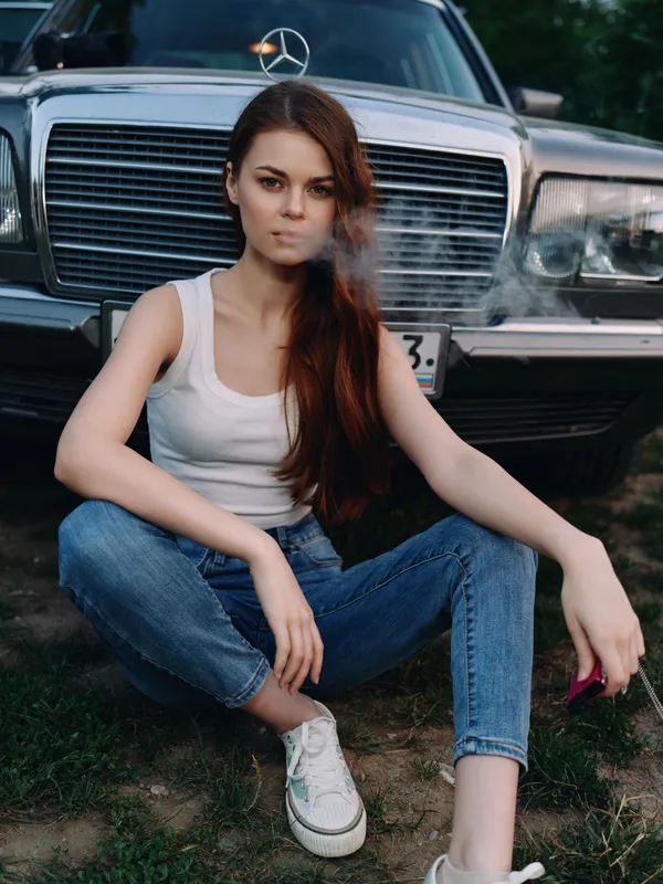 Young woman with long hair sitting casually in front of a vintage car, exuding a relaxed vibe while smoking, embodying urban style and freedom.
