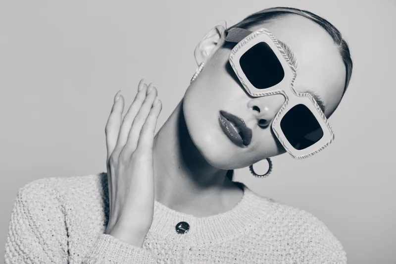 Stylish caucasian woman with fair skin wears oversized sunglasses and light sweater posing with hand near face in monochrome studio portrait.