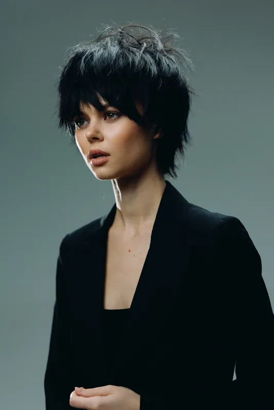 Young woman with short black hair wearing a dark blazer posing against a gray background.